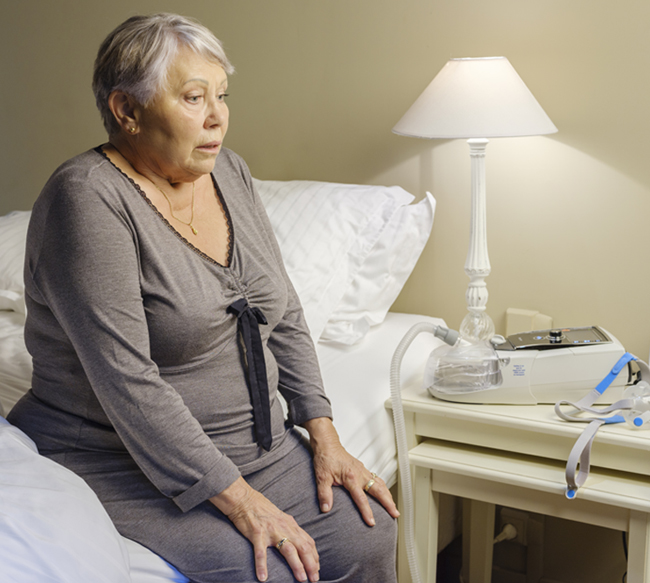 COPD-Patientin sitzt auf dem Bett mit Beatmungsgerät auf dem Nachttisch.