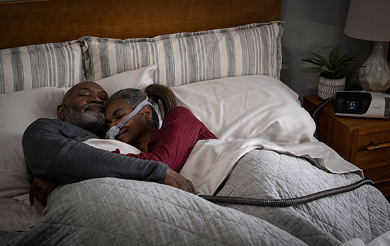 Paar liegt schlafend sich umarmend im Bett. Die frau trägt eine ResMed CPAP-Maske und nutzt das AirSense 11 PAP-Gerät